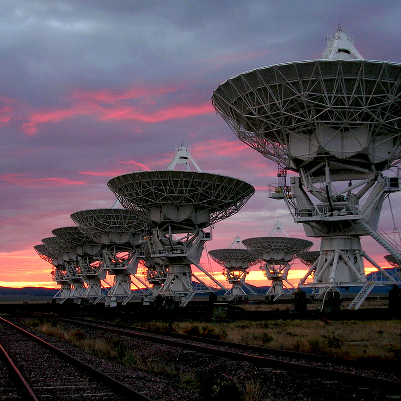 Dramatic photo of an array of radiotelescopes backlit by a sunset glowing orange, purple, and pink.